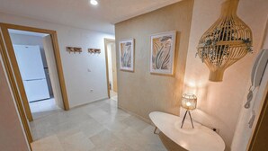 Un couloir avec un sol blanc, une table ronde blanche surmontée d’une lampe, et deux œuvres d’art encadrées accrochées au mur.