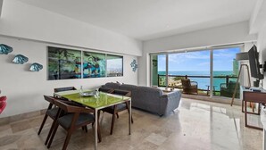 A spacious living area with a dining table, a gray sofa, and a large window offering a view of the ocean.