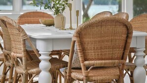 Un comedor con sillas de mimbre y una mesa blanca, ventanas amplias con vistas al exterior y una lámpara colgante.