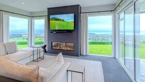A modern living room with a large flat-screen TV mounted on a dark wall, a fireplace, a sofa, and large windows offering a view of a green landscape.