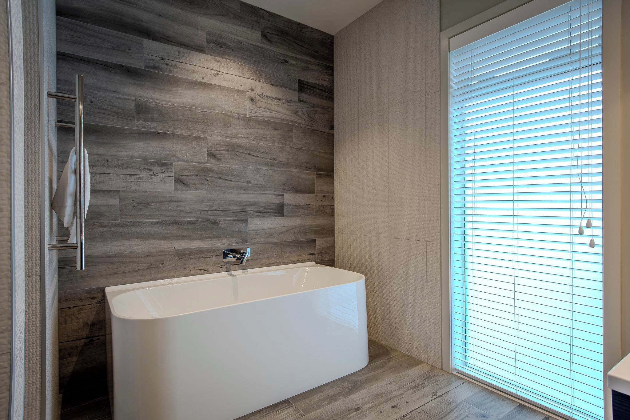 A modern bathroom with a freestanding bathtub, wooden wall paneling, and a large window with blinds.