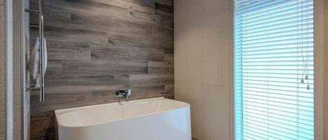 A modern bathroom with a freestanding bathtub, wooden wall paneling, and a large window with blinds.
