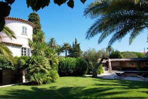 A well-maintained garden with a white building, palm trees, and a thatched-roof structure. - Saint-Tropez center on foot – Villa 5★, 5 suites, pool & garden – 10 pers. (Saint-Tropez)