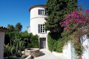 Un bâtiment blanc doté d’une tour ronde, d’une fontaine en pierre et d’une végétation luxuriante comprenant des cactus et des plantes à fleurs.