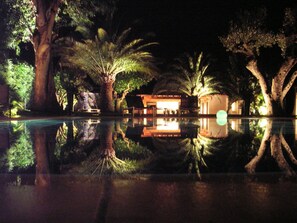 A nighttime view of a resort with a pool reflecting palm trees and a building with warm lighting.