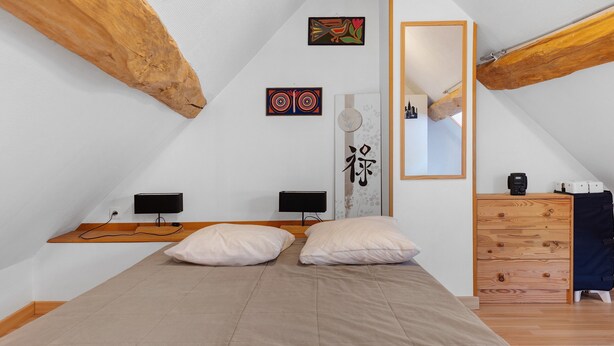 Ein Schlafzimmer mit schrägen Wänden, einem Bett mit zwei Kissen, einer Kommode mit Spiegel und Bildern an der Wand.