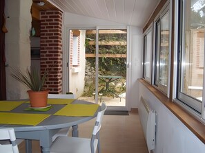 A room with a table, chairs, a potted plant, a sliding glass door leading outside, and a window with blinds. - Gîte Mélany for 4 Guests (Chargé)