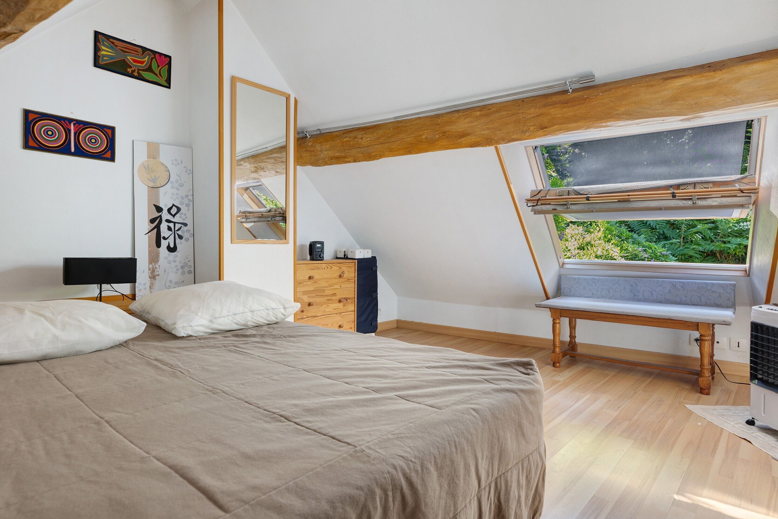 Een slaapkamer met een schuine kap, een bed, een stoel, een bureau en een raam met uitzicht op groen.