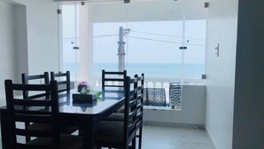 Un comedor con mesa y sillas, una ventana con vista al mar y un balcón con barandilla.