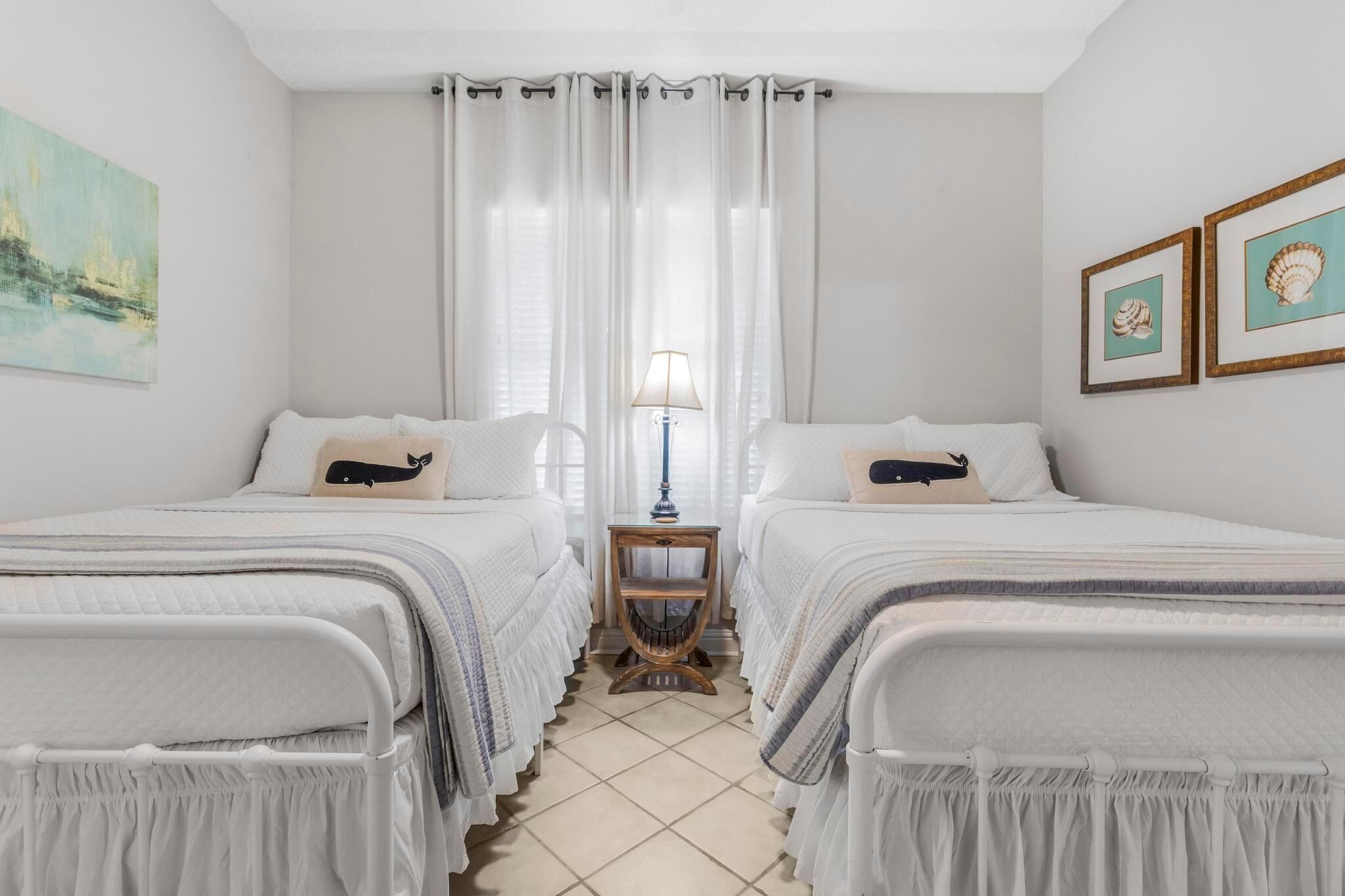 A room with two beds, a nightstand with a lamp, and a window with curtains.