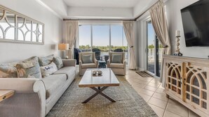 Amplia sala de estar con sofá, mesa de centro y televisor. Balcón con sillas y vista a zonas verdes.