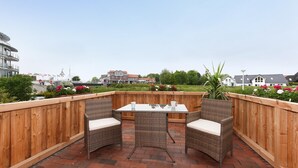 A wooden deck with a table and two chairs, surrounded by a wooden fence, overlooking a residential area with houses and greenery. - Ferienwohnung in Bensersiel, 300 m vom Strand (Bensersiel)