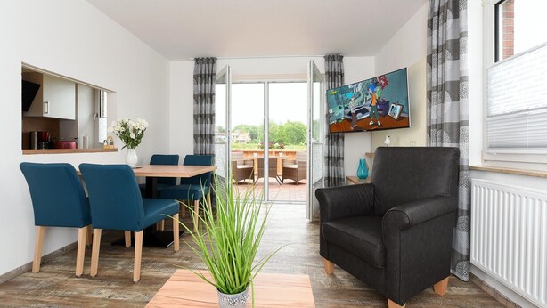 A modern living room with a dining area, a black leather sofa, and a large window with curtains. - Ferienwohnung in Bensersiel, 300 m vom Strand (Bensersiel)