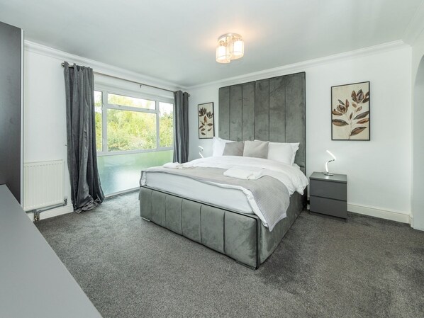 A modern bedroom with a large bed, grey curtains, and a view of greenery outside.