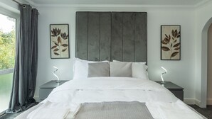 Uma cama bem arrumada com cabeceira cinza, duas mesas de cabeceira com luminárias e dois quadros com folhas nas paredes.