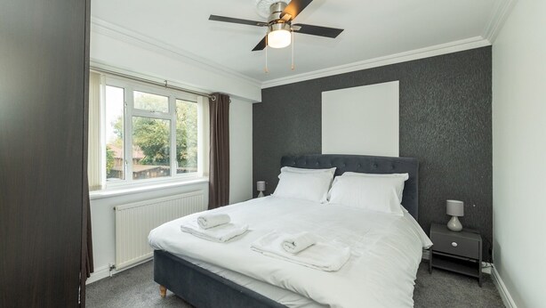 A bedroom with a dark grey bed, white linens, a ceiling fan, a window with curtains, and a radiator. - Pass the Keys | Bright House with Smart TV & Home Comforts (Reading)