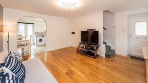 A spacious living area with hardwood flooring, a flat-screen TV on a wooden stand, a dining area with a table and chairs, and a kitchen visible through an archway. - Pass the Keys | Bright House with Smart TV & Home Comforts (Reading)