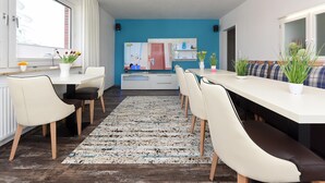 A modern kitchen with a blue accent wall, a dining area with white chairs and a long table, and a large window with curtains. - Ferienwohnung mittendrin in Bensersiel (Bensersiel)