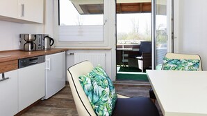 A compact kitchen with a coffee maker, kettle, and dishwasher. There is a dining area with a table and chairs, and a view of the outside through a sliding glass door. - Ferienwohnung mittendrin in Bensersiel (Bensersiel)