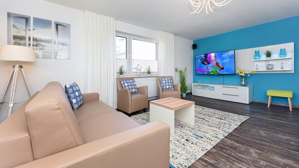 A modern living room with a beige sofa, two armchairs, a coffee table, a TV, and a blue accent wall. - Ferienwohnung mittendrin in Bensersiel (Bensersiel)
