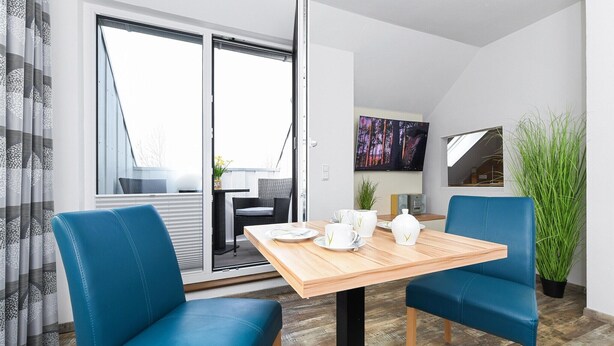 Ein moderner Essbereich mit einem Holztisch, zwei blauen Stühlen, einem Fernseher an der Wand und einer großen Pflanze.