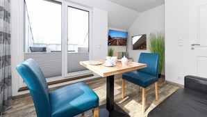 Ein modernes Wohnzimmer mit Holztisch, blauen Stühlen, einem grauen Sofa und einem großen Fenster mit Jalousien.