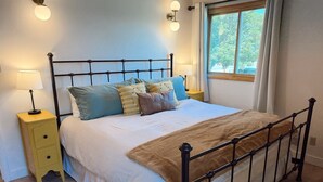 A bed with a metal frame, a bedside table, and a window with curtains. - Main Street Retreat - Beautiful Home (Miles City)
