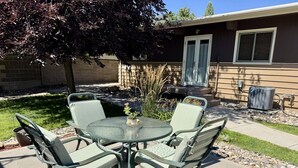 A patio with a round glass table and four chairs, a tree, and a building in the background. - Main Street Retreat - Beautiful Home (Miles City)