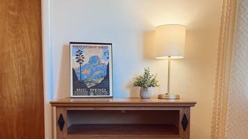 Une commode en bois avec des tiroirs, une affiche accrochée au mur au-dessus, une lampe et une porte visible au fond.