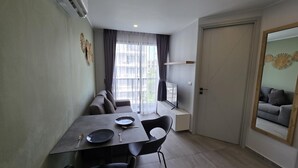 A modern living room with a sofa, a dining table with chairs, a TV on a stand, and a door leading to another room. - Stylish Urban Escape 1Bed Apt T2-822 (Chalong)