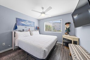 Une chambre à coucher comprenant un lit, un bureau, une chaise, un ventilateur de plafond, une télévision et une fenêtre avec des stores.