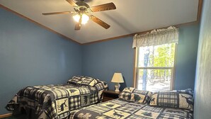 Una habitación con dos camas, un ventilador de techo, una ventana con vista a árboles y una lámpara en una mesita de noche.