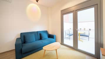 Una sala moderna con un sofá azul, una mesa de centro de madera y un amplio ventanal corredizo que da a un espacio exterior con mesa y sillas.