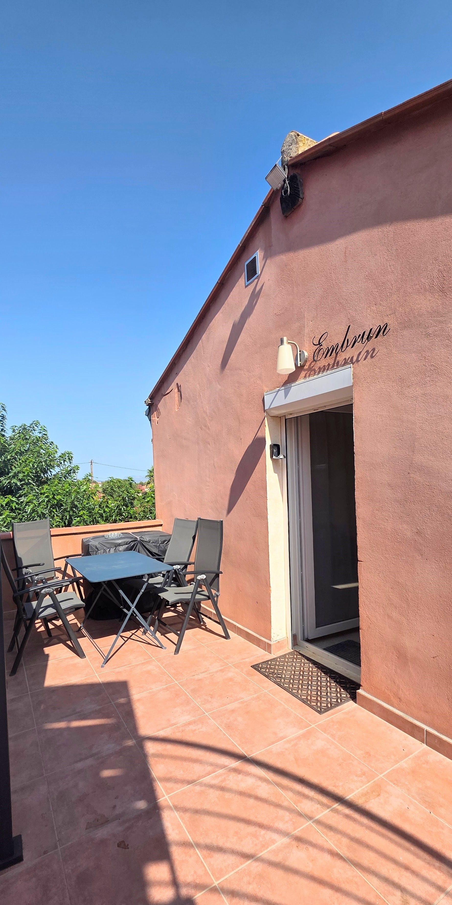 Une terrasse extérieure avec une table et des chaises, une porte et un mur sur lequel est affiché un panneau.