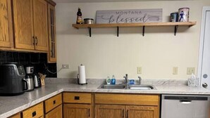 A kitchen with wooden cabinets, a dishwasher, a coffee maker, and a sign above the sink that reads "MONTANA blessed." - Studio House - Central Location (Miles City)
