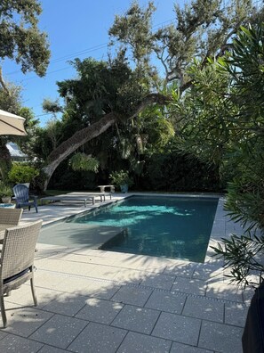 Une piscine rectangulaire, bordée d’une terrasse carrelée et entourée d’une végétation luxuriante et d’arbres.