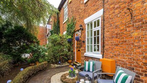 A brick building with a small garden area featuring a seating arrangement with a table and chairs, a small circular table with a plant, and a pathway leading to a door. - Beautiful Cozy Cottage Home in Sandbach, Sleeps 4! (Cheshire East)