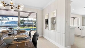A dining area with a wooden table set for four, a large window with a view of a swimming pool, and white paneled walls. - European Artistic Retreat (Cape Coral)