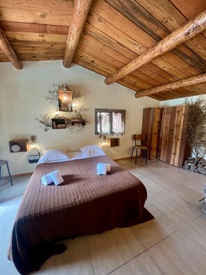 Une chambre avec un plafond en bois, un lit recouvert d’une couverture marron, deux tables de chevet avec des lampes, une fenêtre avec des rideaux et une petite plante.