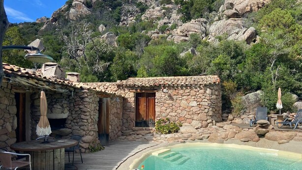 A stone building with a pool, outdoor seating, and a mountainous backdrop.