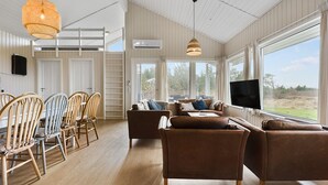A spacious living area with a wooden floor, a brown leather sofa set, a dining table with chairs, and large windows offering a view of the outdoors. - Cozy home in Løgstør with sauna (Løgstør)