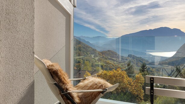A chair with a fur cushion, a glass window, and a view of a valley with mountains. - Eco del Garda – Room 306 – with Covered Terrace and Lake View (Tenno)