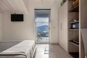 A bedroom with a bed, a TV mounted on the wall, a sliding door leading to a balcony with a view of mountains, and a wooden cabinet with shelves.