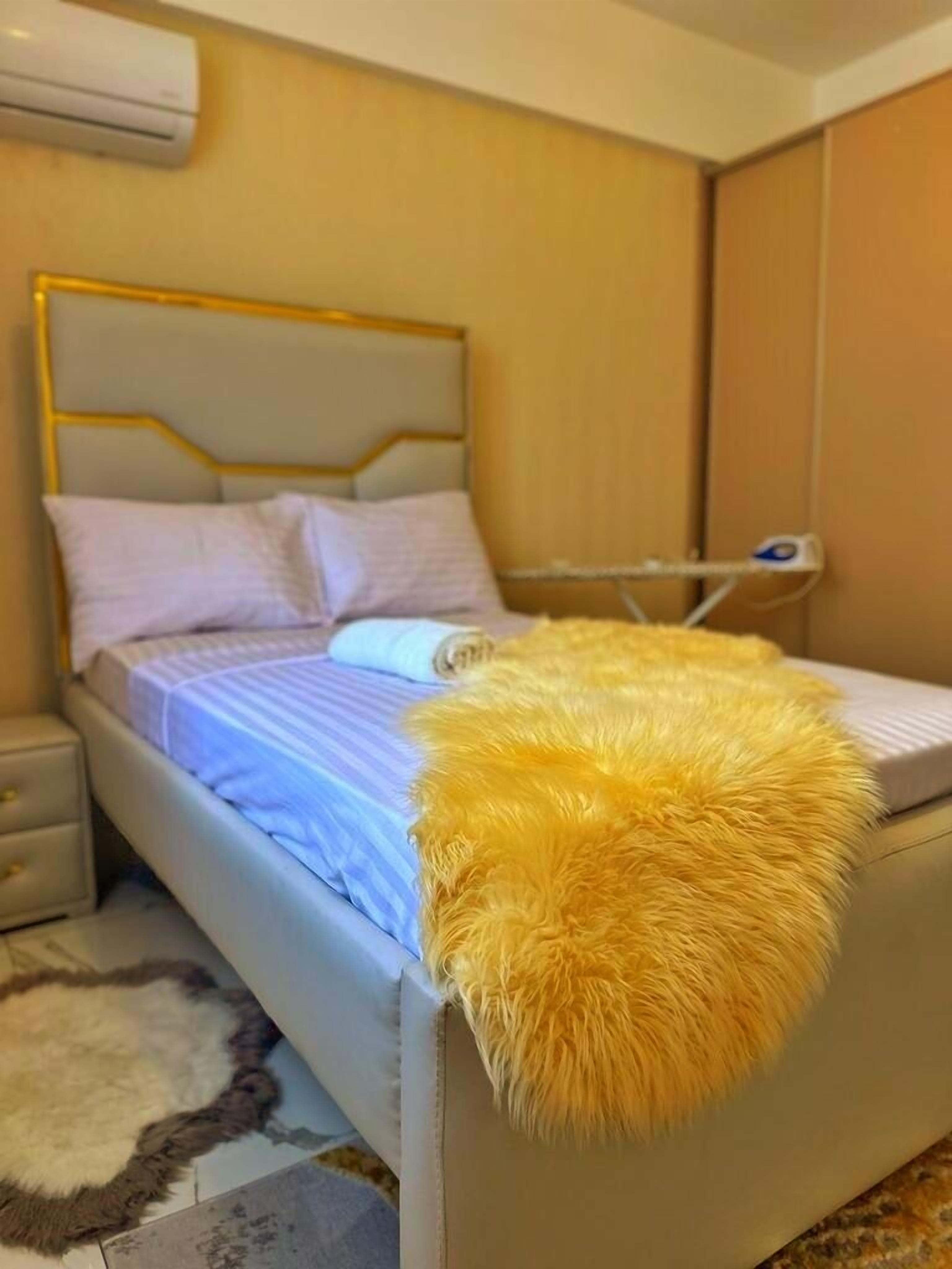 A bedroom with a bed, a bedside table, a lamp, and a fluffy yellow pillow.