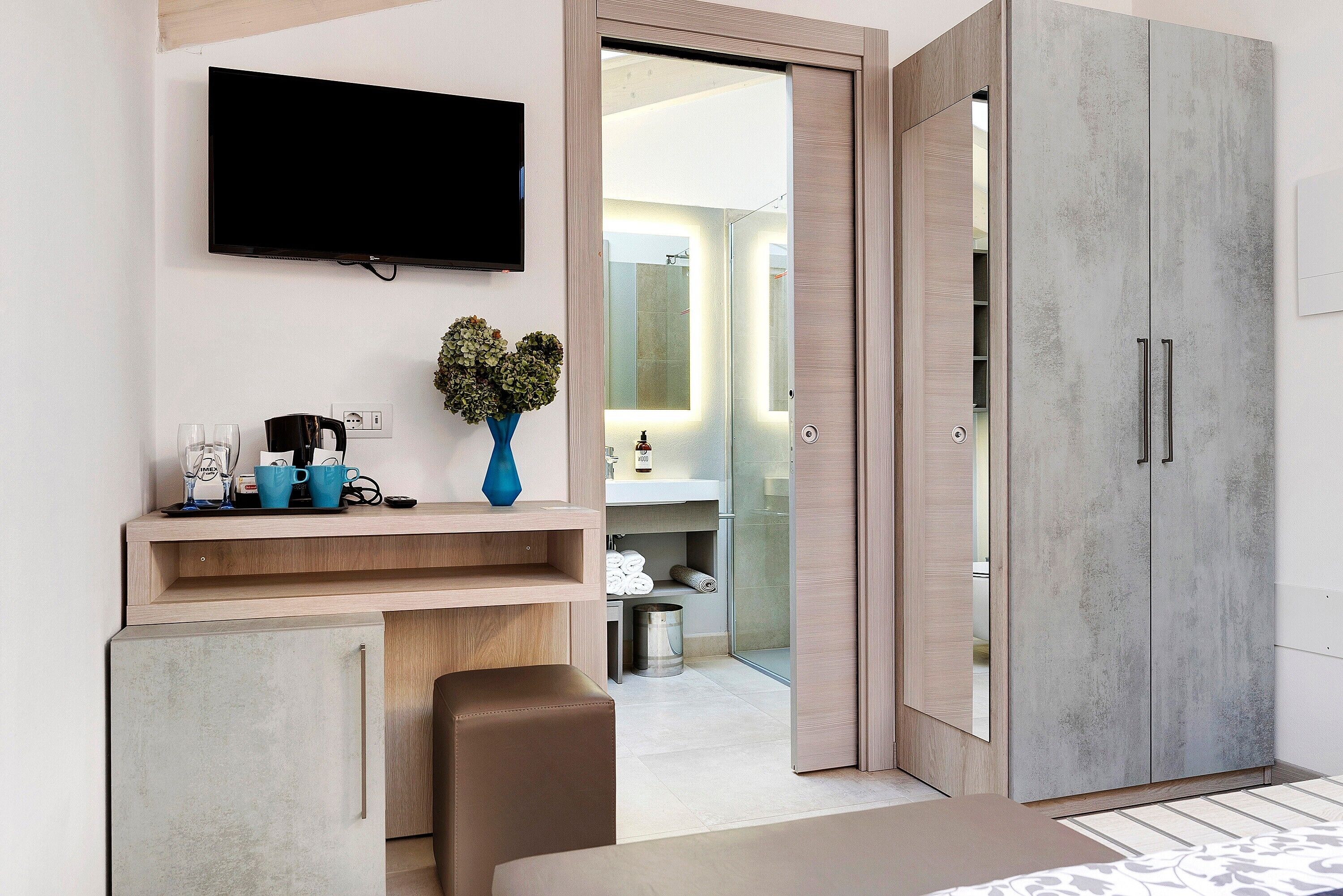 A modern hotel room with a flat-screen TV, a small desk with a coffee maker, a vase with greenery, and a wardrobe with a mirror.