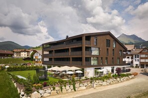 Ein modernes Mehrstöckiges Hotel mit Außenbestuhlung, einer Steinsichtmauer und einer Bergkulisse.
