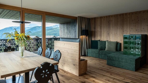 A modern living room with a wooden floor, a dining table with chairs, a sofa, a green cabinet, and a view of mountains through large windows.