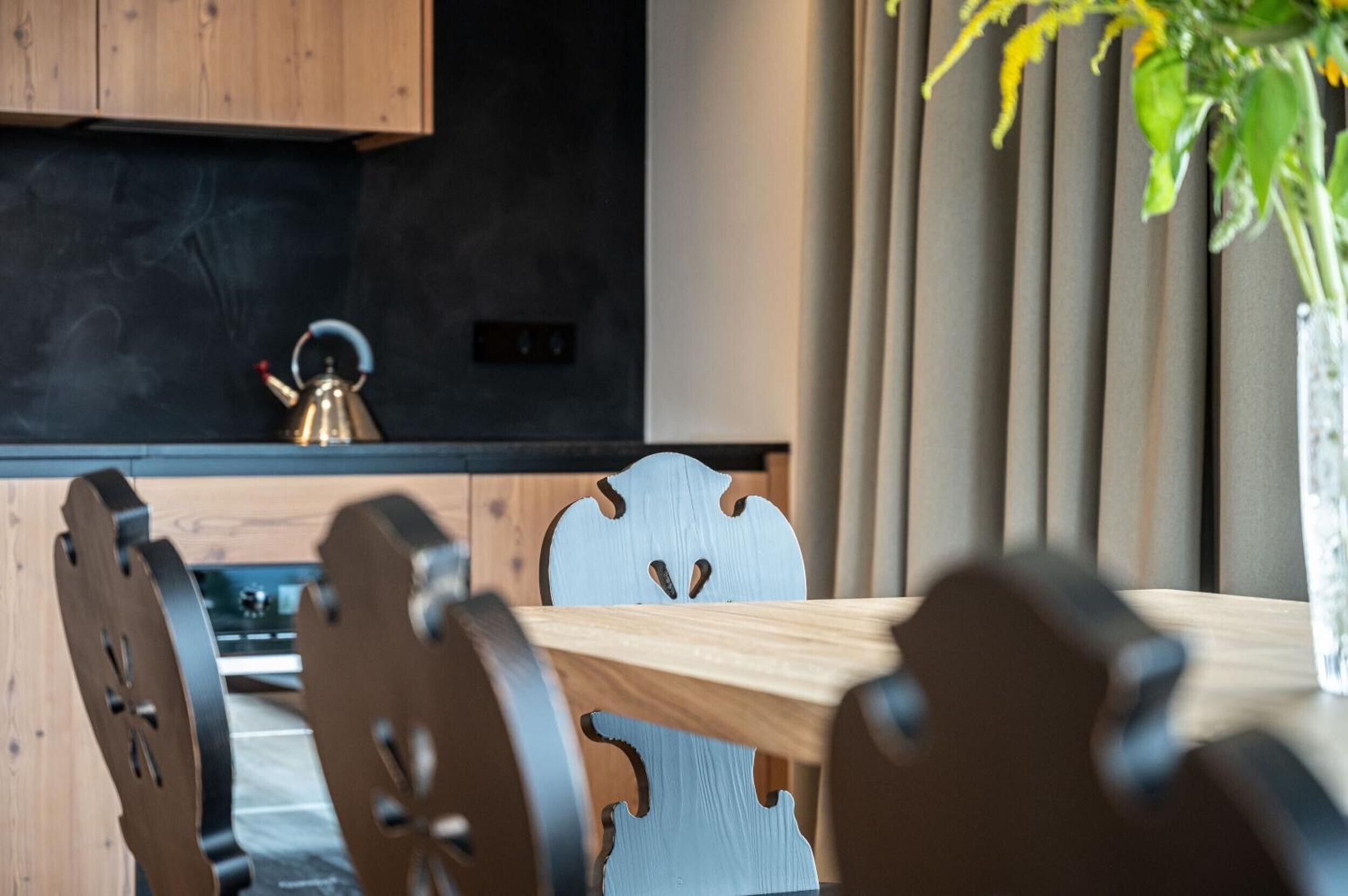 A modern kitchen with a wooden table and chairs, a kettle, and a plant in a vase.