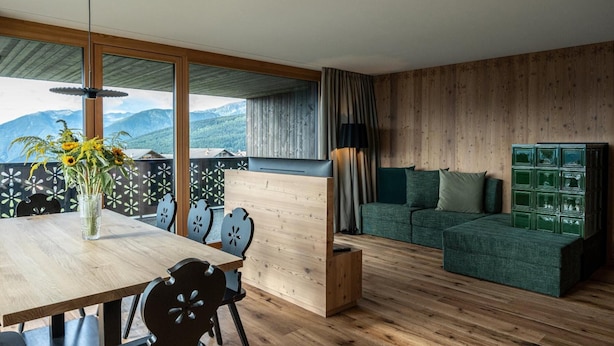 A modern living room with a wooden floor, a dining table with chairs, a sofa, a green cabinet, and a view of mountains through large windows.