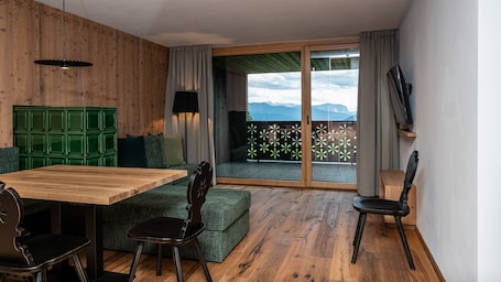 A room with a wooden floor, a wooden table, a green sofa, a wooden chair, a TV on the wall, and a view of mountains through a glass door.